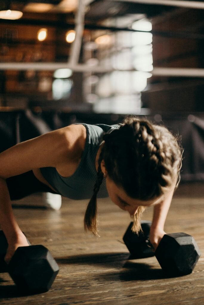 Adeus, Mensalidades! Descubra por que Milhares de Pessoas trocaram a Academia pelo Treino em Casa. Fit woman doing push-ups with dumbbells indoors, emphasizing strength training and fitness.